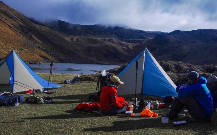 Two tents sit in an open area beside an alpine lake. There are mountains surrounding the lake.